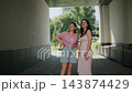 Two women stand under a covered walkway, looking outward. One wears a pink top and shorts, smiling. The other wears a pink dress, arms crossed, expression neutral 143874429