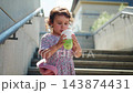 Child sips drink while walking down urban concrete stairs. She holds cup with both hands, focused on beverage. Natural light highlights her curly hair and floral dress. Scene captures casual 143874431