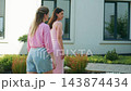 Two women in pink outfits stand outdoors near a modern house. One raises her arm while both gaze upward with curiosity. Bright sunlight enhances their casual, cheerful mood 143874434