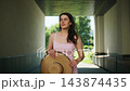 Woman in pink dress holds straw hat. She stands in shaded walkway with greenery beyond. She adjusts hat, eyes closed, calm and relaxed. Footage ideal for summer, travel, or fashion themes 143874435