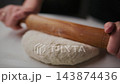 Hands roll dough with wooden roller on white surface. Action shows dough flattening slowly. Focus on rolling motion and texture of dough. Ideal for cooking, baking, or food preparation content 143874436