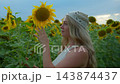 Woman with long blonde hair gently touches a sunflower. She wears a light-colored knit cap. Background shows vast sunflower field under soft evening sky. Scene evokes calm, peaceful 143874437