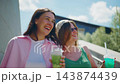 Two young women smile while holding colorful drinks outdoors. Bright sunny day highlights their joyful expressions. They walk on modern architecture steps. Footage suits social events, travel 143874439