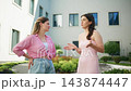 Two women argue outside modern building. One has hands on hips, looking frustrated. Other gestures with hands, speaking passionately. Scene shows emotional conflict during daytime 143874447