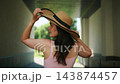 Woman adjusts wide-brimmed hat under shaded walkway. Sunlight filters through trees behind her. She wears light pink sleeveless dress. Appears relaxed and confident while walking forward 143874457