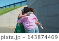 Two women embrace warmly outdoors near stone walls. One wears green, other pink. They hold drinks, smile gently. Child stands nearby, watching. Scene captures joyful friendship and casual meet-up 143874469