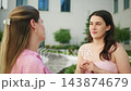 Two women smile and gesture while chatting outside. They appear joyful and engaged in friendly conversation. Scene suggests casual, warm social interaction. Ideal for themes like friendship 143874679