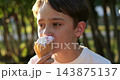 Teenager eats ice cream cone outdoors in sunny park. Young male enjoys sweet summer treat in warm sunlight. Close-up captures happy expression, carefree youth, relaxation and outdoor leisure. 143875137