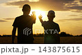 Father and son raise hands for high five in open field at sunset. Silhouettes stand together against golden sky and warm sunlight. Scene represents family bonding, support, childhood and togetherness. 143875615