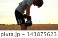 Young boxer rests bent forward in harvested field at sunset. Boy wearing boxing gloves recovers after outdoor workout. Warm sunlight creates calm mood for resilience and fitness stories. 143875623