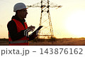 Power line inspector reviews data on tablet at sunset. Utility engineer checks transmission tower in field. Industrial safety and energy infrastructure inspection supports reliable grid operations. 143876162