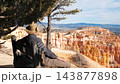 Male tourist wearing cowboy hat sitting on rustic wooden bench under trees, admiring colorful sandstone hoodoos and natural orange rock formations in bryce canyon national park in utah state 143877898