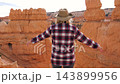 Sporty female hiker wearing hat standing on top cliff edge, raising her arms in triumph while looking at red orange rock hoodoos during mountain journey. Joy of successfully reaching the top trail 143899956