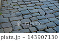 Old Uneven Cobblestone Street with Metal Drain Grate and Weathered Stone Pavement in Historic Urban Area 143907130