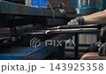 Workers sit side by side, fitting knitted protective gloves onto metal forms moving along conveyor. Repetitive manual loading keeps industrial glove production running at steady pace. 143925358
