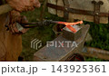 Macro closeup shot captures strong hands of medieval blacksmith hammering a glowing red-hot steel workpiece held with tongs on a heavy iron anvil in traditional outdoor forge... 143925361