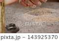 Closeup macro of dusty, flour-covered hand feeding and guiding crushed wheat grains into the central eye hopper of a traditional granite quern stone hand mill. 143925370