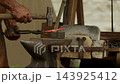 Medium closeup shot captures medieval blacksmith striking a glowing red-hot steel bar with a heavy hammer on an iron anvil beside a portable stone forge hearth in an open-air workshop... 143925412