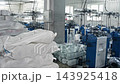 Wide shot reveals busy textile factory floor where automated blue knitting machines produce garments. Large white sacks filled with finished products sit piled near concrete pillar. 143925418