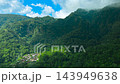 Aerial view of dense green forested mountain slopes, cloudy peaks, and a remote settlement nestled 143949638
