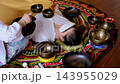 High angle view of a therapist playing a Tibetan singing bowl near a woman resting with an eye pillow. Private sound bath rituals support stress relief, deep relaxation, and emotional balance. 143955029